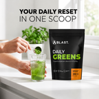 Person adding Blast Daily Greens powder to a glass with a BLAST Daily Greens package on a white surface.