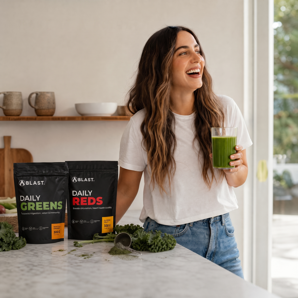 A woman in a white t-shirt and jeans stands in a bright kitchen holding a glass of green juice, smiling. In the foreground on a marble counter are two black BLAST supplement pouches labeled Daily Greens and Daily Reds, with kale scattered nearby.