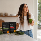 A woman in a white t-shirt and jeans stands in a bright kitchen holding a glass of green juice, smiling. In the foreground on a marble counter are two black BLAST supplement pouches labeled Daily Greens and Daily Reds, with kale scattered nearby.