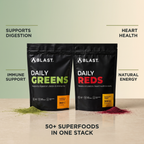 Two black BLAST supplement pouches on a beige surface with text callouts around them: 'SUPPORTS DIGESTION' next to Daily Greens, 'IMMUNE SUPPORT' on the left, 'HEART HEALTH' on the right, and 'NATURAL ENERGY' on the right. The left pouch is labeled Daily Greens with green text and a small orange flavor block; the right pouch is labeled Daily Reds with red text and a small yellow flavor block.