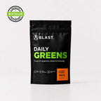 A black stand-up pouch of BLAST Daily Greens supplement powder on a light gray background, with a green and black Bestseller badge in the top-left corner. The pouch features white and bright green text, and an orange label indicating Mango & Orange flavor and 300g weight.