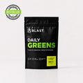 A black stand-up pouch of BLAST Daily Greens supplement on a light gray background, with a green 'BESTSELLER' badge in the top-left corner. The pouch features white and bright green text, and a yellow label indicating pineapple and lemon flavor, 300 g.