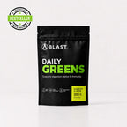 A black stand-up pouch of BLAST Daily Greens supplement on a light gray background, with a green 'BESTSELLER' badge in the top-left corner. The pouch features white and bright green text, and a yellow label indicating pineapple and lemon flavor, 300 g.