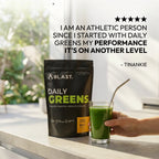 A black stand-up pouch of BLAST. Daily Greens powder sits on a countertop next to a hand holding a glass of green drink with a metal straw.